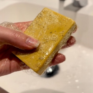 May include: A yellow bar of soap with a textured surface is being held in a hand over a white sink. The soap is covered in white bubbles.