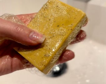 May include: A yellow bar of soap with a textured surface is being held in a hand over a white sink. The soap is covered in white bubbles.