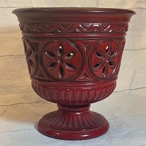 May include: A dark red, carved wooden urn with a pedestal base. The urn features decorative floral cutouts and geometric patterns. The vessel has a wide rim and a detailed, textured surface. The overall design is ornate and elegant.