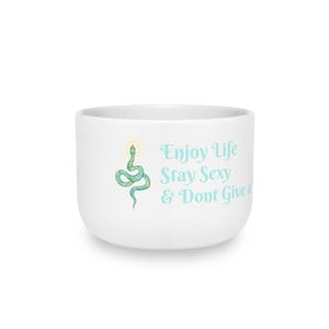 May include: A white ceramic mug featuring a teal snake illustration with a yellow halo above its head. To the right of the snake, the text reads: Enjoy Life Stay Sexy & Dont Give a. The design is printed in a light teal colour on the smooth surface.