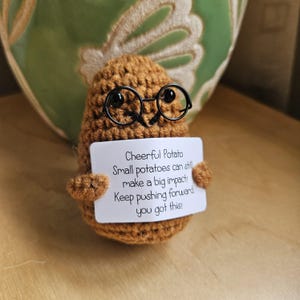 May include: A handmade, crocheted potato in shades of brown, wearing small black spectacles. It holds a white sign with the text: "Cheerful Potato, Small potatoes can still make a big impact! Keep pushing forward, you got this!"