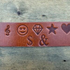 May include: A brown leather strap with various symbols engraved on it, including a musical note, a smiley face, a diamond, a star, a heart, a dollar sign, and an ampersand.