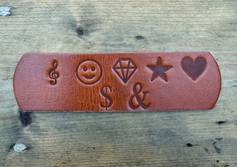 May include: A brown leather strap with various symbols engraved on it, including a musical note, a smiley face, a diamond, a star, a heart, a dollar sign, and an ampersand.