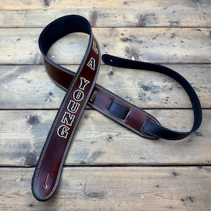 Peut inclure: Une sangle de guitare en cuir marron avec le nom "L A YOUNG" en lettres blanches. La sangle a un dos noir et des coutures blanches. La sangle mesure environ 6,35 cm de large et 127 cm de long.