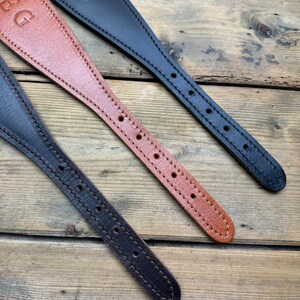 May include: Three leather dog collars in different colors. The collars are brown, black, and tan. Each collar has a name engraved on it. The collars are laid out on a wooden surface.