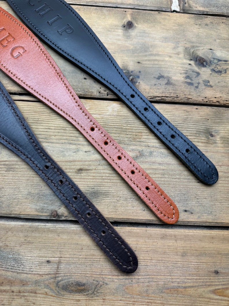 May include: Three leather dog collars in different colors. The collars are brown, black, and tan. Each collar has a name engraved on it. The collars are laid out on a wooden surface.