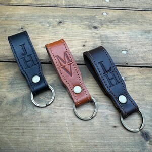 May include: Three leather keychains with metal rings. The keychains are brown, black, and dark brown. Each keychain has a different monogram embossed on it: "JH", "MV", and "LH".
