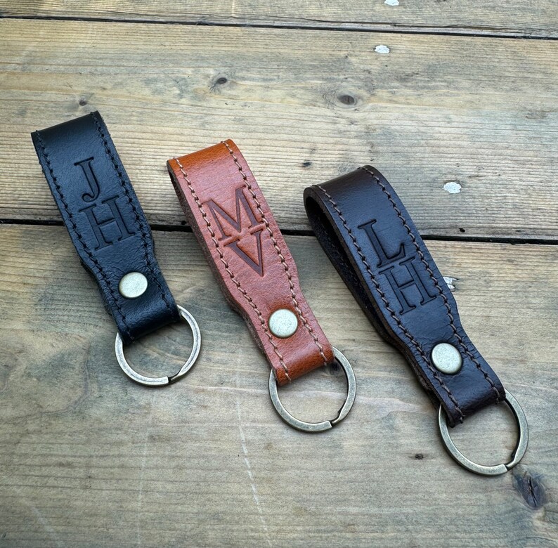 May include: Three leather keychains with metal rings. The keychains are brown, black, and dark brown. Each keychain has a different monogram embossed on it: "JH", "MV", and "LH".