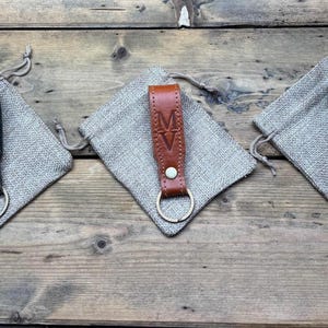 May include: Three leather keychains with a metal ring. Each keychain has a different initial engraved on it. The keychains are displayed on a burlap surface.