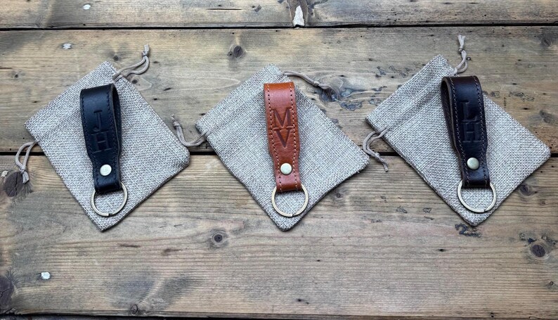May include: Three leather keychains with a metal ring. Each keychain has a different initial engraved on it. The keychains are displayed on a burlap surface.