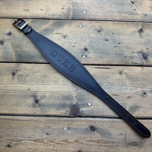 May include: A brown leather dog collar with a brass buckle and the name "DUKE" embossed on the front.