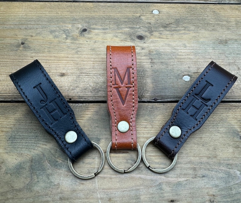 May include: Three leather keychains with metal rings. The keychains are brown, black, and dark brown. Each keychain has a different set of initials embossed on the leather. The initials are "JH", "MV", and "JH".