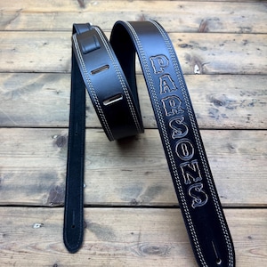 Op de afbeelding: Zwarte leren gitaarband met witte stiksels en de naam "PARSONS" in zilveren letters gegraveerd.