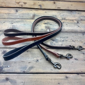 Könnte beinhalten: Drei Hundeleinen aus Leder in Schwarz, Braun und Beige, jeweils mit kontrastierenden weißen Nähten und einem Metallverschluss. Die Leinen liegen auf einer Holzoberfläche und zeigen ihr Design.