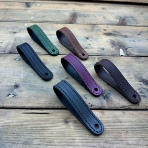 May include: Six leather loops in different colours, including green, brown, burgundy, black, and dark grey. The loops are stitched with a contrasting thread and have a hole at the end.