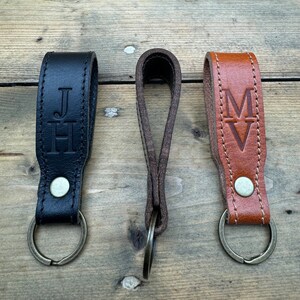 May include: Three leather keychains with metal rings. The keychain on the left is black with the initials "JH" embossed on it. The keychain in the middle is brown with no personalization. The keychain on the right is brown with the initials "MV" embossed on it.
