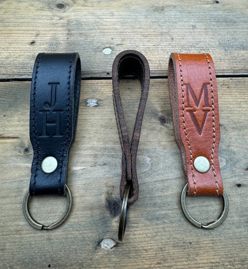 May include: Three leather keychains with metal rings. The keychain on the left is black with the initials "JH" embossed on it. The keychain in the middle is brown with no personalization. The keychain on the right is brown with the initials "MV" embossed on it.