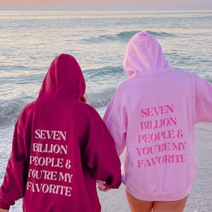 Peut inclure: Deux personnes portant des sweats à capuche rose et bordeaux avec le texte "Seven Billion People & You're My Favorite" au dos. Elles sont debout sur une plage avec la mer en arrière-plan.
