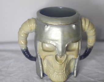 May include: A ceramic mug shaped like a skull wearing a silver helmet with curved horn handles. The skull is off-white with a detailed jawline, and the interior is dark blue.