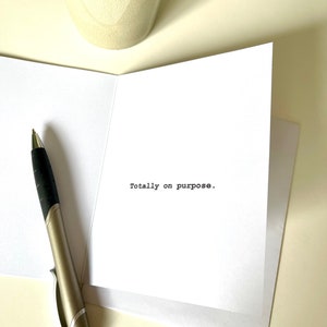 May include: A white card with the text "Totally on purpose." printed in black typewriter font. A silver pen is lying on the card.