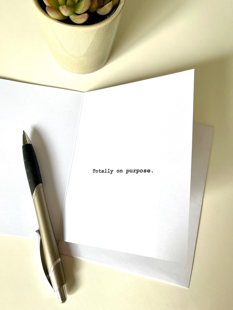 May include: A white card with the text "Totally on purpose." printed in black typewriter font. A silver pen is lying on the card.