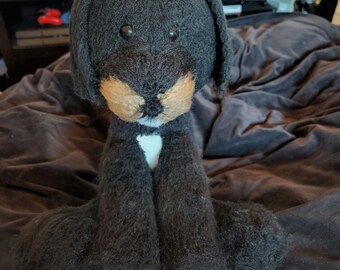 Puede incluir: Un juguete de peluche de perro negro y marrón con marcas blancas en el pecho. El juguete está sentado en una manta marrón.