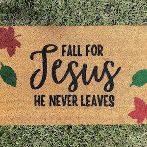 May include: A brown coir doormat with the text "Fall for Jesus He Never Leaves" in black lettering. The doormat is decorated with red and green leaf designs.
