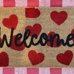 May include: A brown coir doormat with the word "Welcome" in black script. The doormat is decorated with a pattern of red hearts.