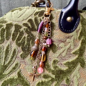 May include: A gold-toned keychain charm with a dragonfly pendant and a cluster of colorful beads. The beads are various shapes and colors, including purple, brown, pink, and yellow. The charm is attached to a gold clasp.