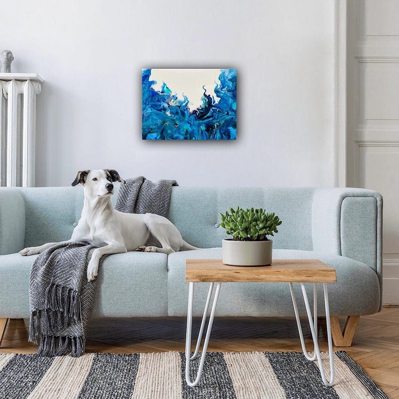 May include: Abstract painting with blue and white colors. The painting is framed and hanging on a white wall above a light blue sofa. A white dog is lying on the sofa. A small potted plant is on a wooden table with white hairpin legs.