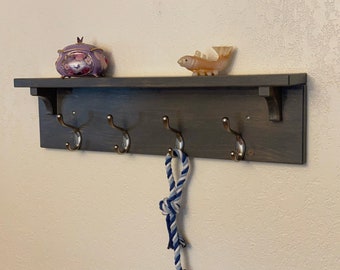 May include: A dark gray wooden shelf with four hooks and a small shelf on top. The shelf is mounted on a white wall. A small purple music box and a small orange fish figurine are on the top shelf.