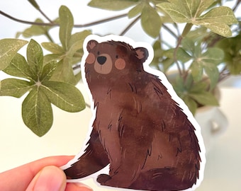 May include: A brown bear sticker with a white border. The bear is sitting with its paws forward, and has a dark brown nose, and rosy cheeks. The sticker is held by a hand in front of a background of green leaves.