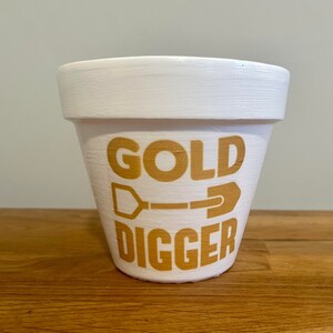 以下が含まれることがあります： 金色の文字で「GOLD DIGGER」と書いてあり、金色のシャベルのグラフィックが描かれた白いセラミック製の植木鉢です。