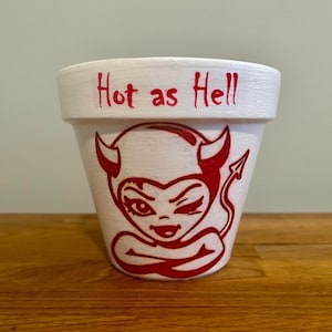 May include: White ceramic flower pot with a red cartoon devil face and the text "Hot as Hell" painted on the side.