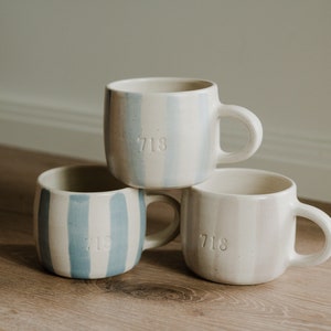 May include: Three ceramic mugs with blue and white striped designs. Each mug has the number "718" embossed on the side.
