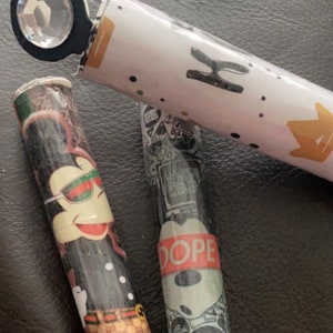 May include: Three colourful tubes with different designs. The tubes are decorated with various patterns, including a cartoon character with sunglasses, a red and white "DOPE" label, and a green and white "TV KILLS EVERYTHING" label.