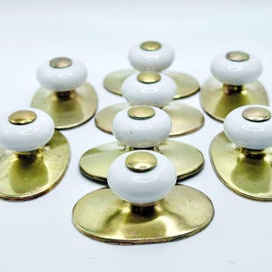 May include: A collection of eight cabinet knobs. Each knob features a white, rounded ceramic top with a small, gold-colored metal disc on top. The knobs are mounted on oval-shaped, gold-colored metal backplates.