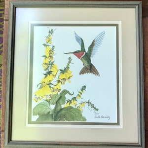 May include: A framed watercolor painting of a hummingbird in flight, with a red throat and green body, hovering near yellow flowers. The painting is signed "Anita Connelly 1995".