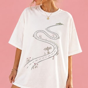 May include: A white short-sleeve t-shirt featuring a black line drawing of a winding road leading to a mountain. The graphic includes a small wooden signpost labeled WRONG and delicate pink flower illustrations scattered along the roadside.