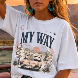 Puede incluir: Una camiseta blanca con un estampado gráfico de un coche descapotable en una carretera desértica. El texto dice MY WAY, con una señal que indica done explaining, better things ahead y no turning back, junto a un cactus.