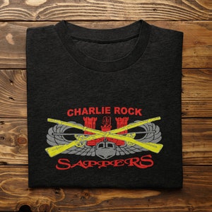 May include: Black t-shirt with a red and yellow graphic of a military insignia. The graphic includes the text "Charlie Rock", "44", and "Sappers".