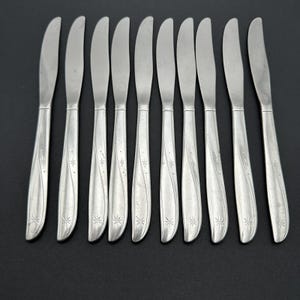 May include: A set of ten stainless steel butter knives arranged on a dark surface. Each knife features a curved handle with a decorative starburst pattern and a smooth, rounded blade. The knives are designed for spreading butter or other soft spreads.
