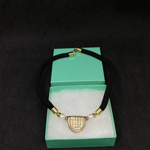 May include: A black braided cord necklace with a gold and silver pendant featuring a cluster of clear rhinestones. The pendant is accented with two white pearls. The necklace is displayed in a light green gift box.