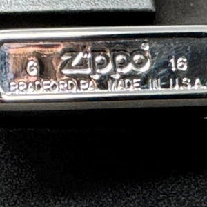 May include: Close-up of a silver Zippo lighter. The base is stamped with the brand name, the number 6, and the text "BRADFORD PA. MADE IN U.S.A." The number 16 is also visible. The lighter is on a black surface.