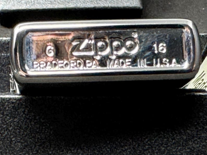 May include: Close-up of a silver Zippo lighter. The base is stamped with the brand name, the number 6, and the text "BRADFORD PA. MADE IN U.S.A." The number 16 is also visible. The lighter is on a black surface.