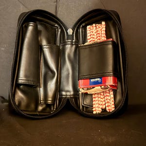 Op de afbeelding: Een zwarte etui met ritssluiting en meerdere binnenvakken. De linkerkant heeft een verticaal vak, terwijl de rechterkant een rood-wit patroonpakje Vauen-filters en een stapel witte en rode pijpenragers bevat.