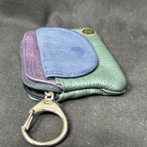 May include: A multi-colored leather key case with a silver-tone carabiner clip. The key case features a purple, blue, and green color scheme. The key case has a zipper closure and a small gold-tone emblem.