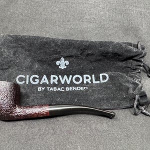 May include: A tobacco pipe with a dark brown, textured bowl and a black stem rests in front of a black drawstring bag. The bag has the text "CIGARWORLD BY TABAC BENDEN" in white.