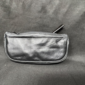 May include: A small, black leather coin purse with a zipper closure and a pull tab. The purse has a rounded shape and a smooth, slightly textured surface. The background is a dark, textured fabric.