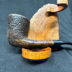 May include: A brown tobacco pipe with a textured bowl and a black stem rests on a wooden stand. The pipe has a curved shape and is made of wood. The stand is also made of wood and has a natural finish.